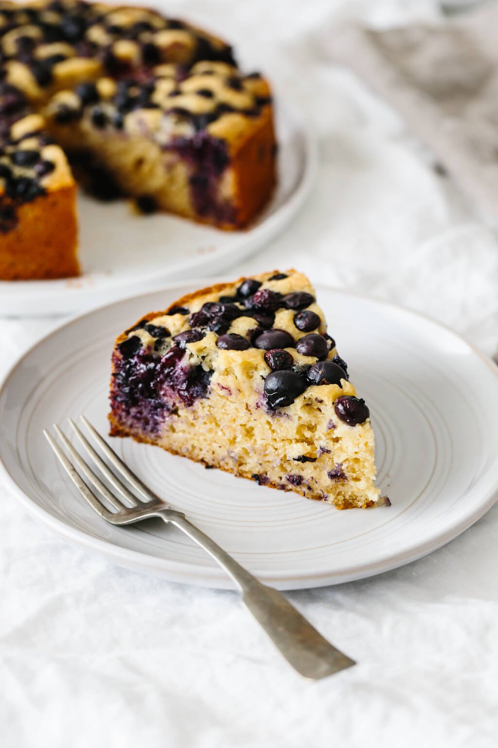 A slice of paleo lemon blueberry cake on a plate, sitting next to the cake.