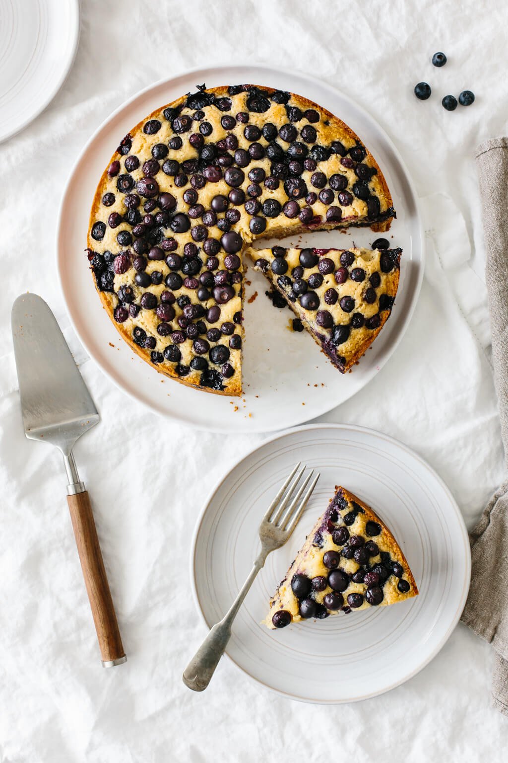A lemon cake topped with blueberries on a table. A slice of the cake is taken from the cake and on a plate next to it.