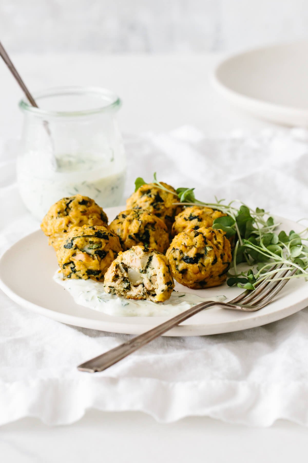 Baked tuna meatballs are a healthy, low-carb, seafood spin on meatballs! A plate of tuna meatballs garnished with tzatziki and micro greens with one half eaten .