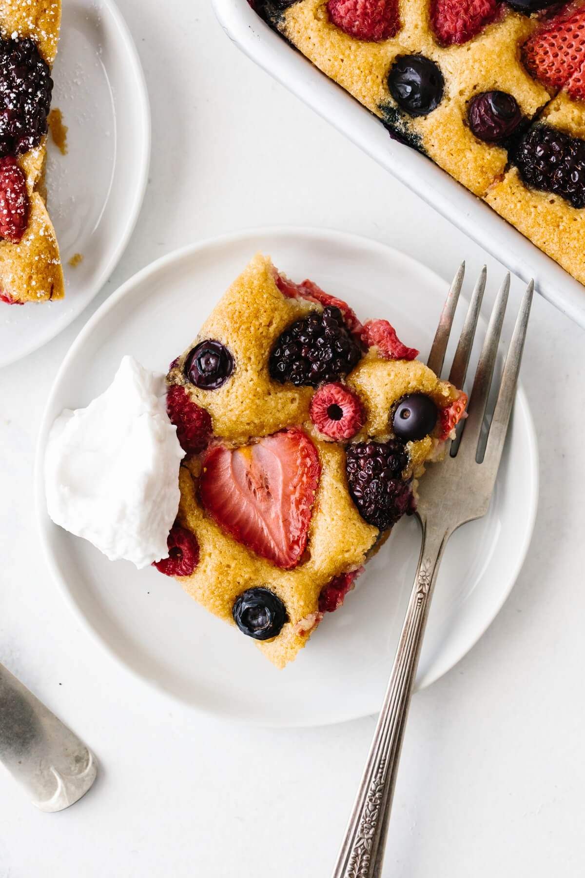 Slice of berry sheet cake on a white plate with fork and dollop of cream.