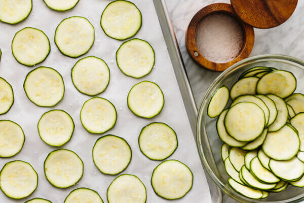 Zucchini slices on top of a parchment lined baking sheet next to a bowl of extra zucchini slices.