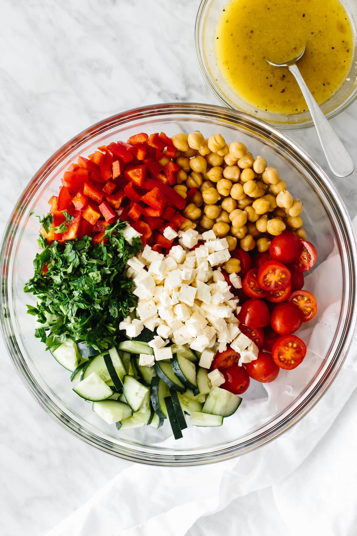 This Mediterranean chickpea salad is the ultimate summer salad that's perfect for all your summer cookouts, dinner parties, and picnics! Ingredients for a Mediterranean chickpea salad in a large glass bowl next to a small bowl of lemon vinaigrette.