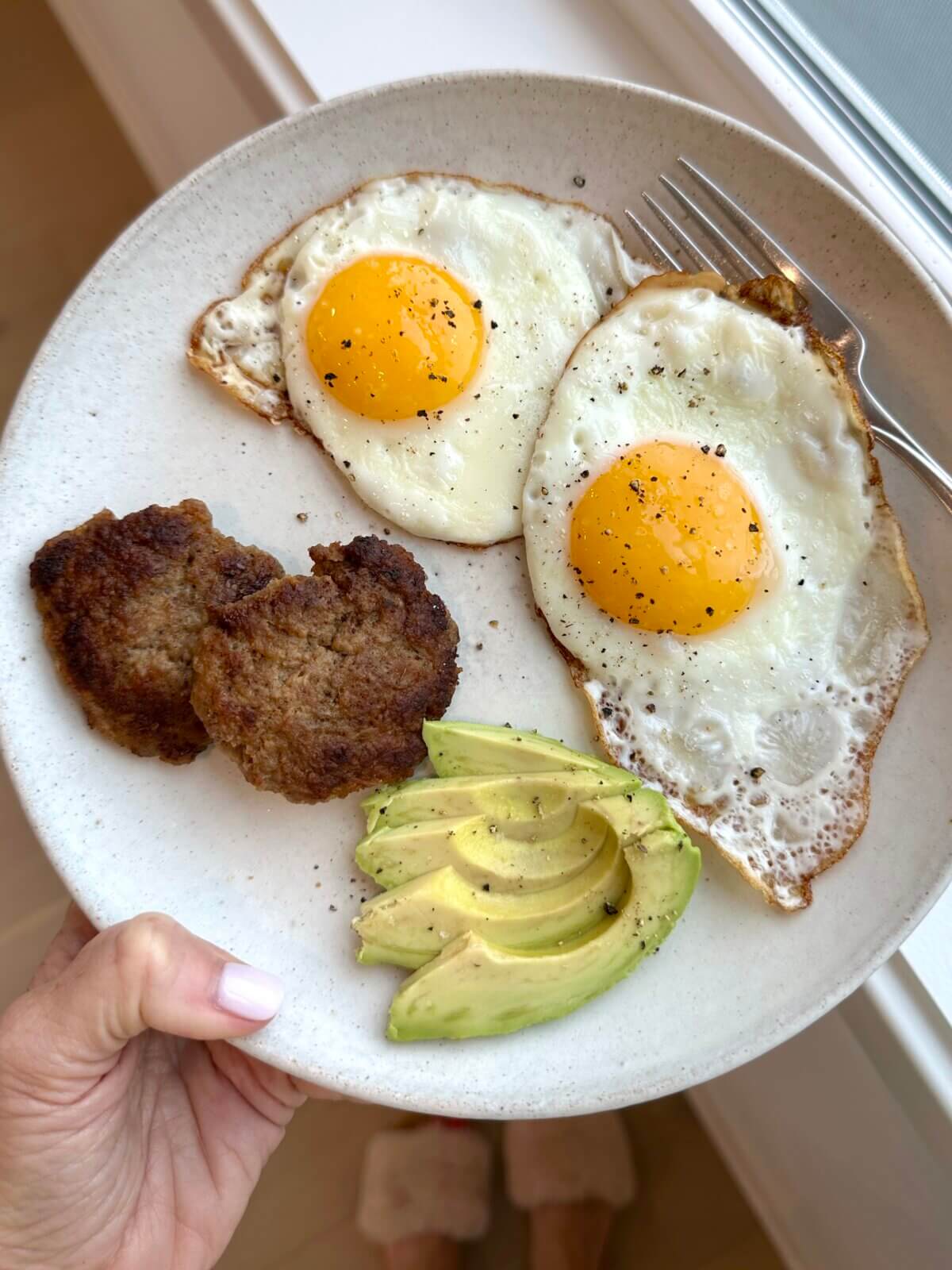 Breakfast sausage, fried eggs, and avocado on a plate.